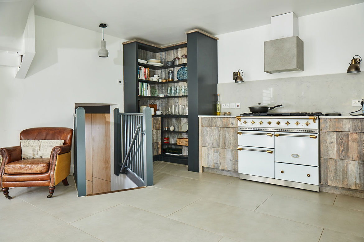 Brown leather armchair sits next to bespoke kitchen with white Lacanche range cooker
