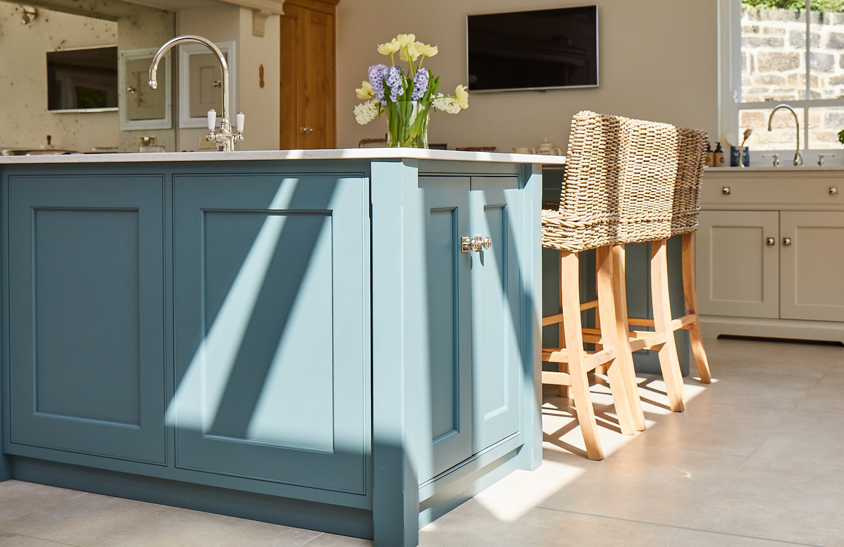 Sun shines on painted green kitchen island with wicker bar stools pushed under worktop