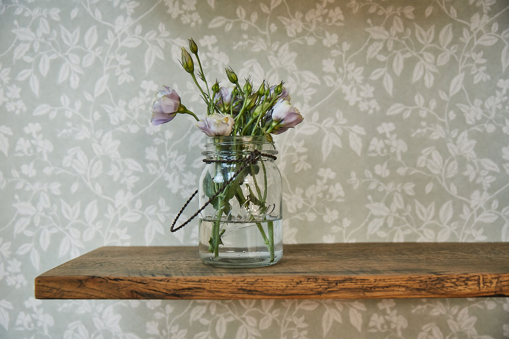Flowers in glass jar sit on rustic oak shelf with green floral wallpaper background