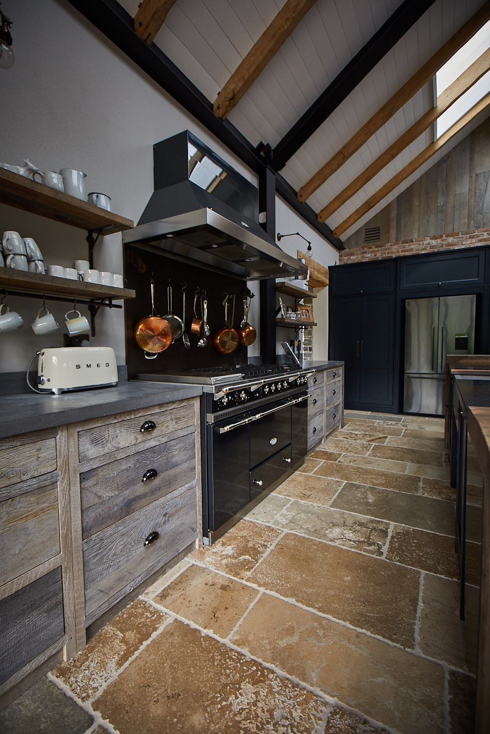Black range cooker with matching hood next to rustic oak pan drawers and rustic open shelves