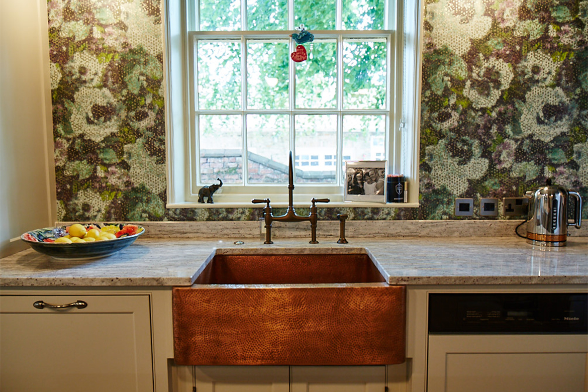 Beaten copper belfast sink with antique copper bridge taps set in front of window with green floral wallpaper
