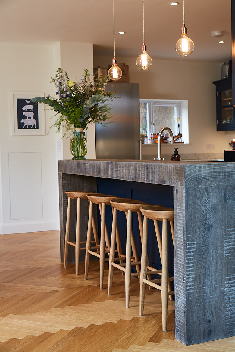 4 clean oak bar stools sit on parquet wood floor under hand aged chunky breakfast bar with blue back panel