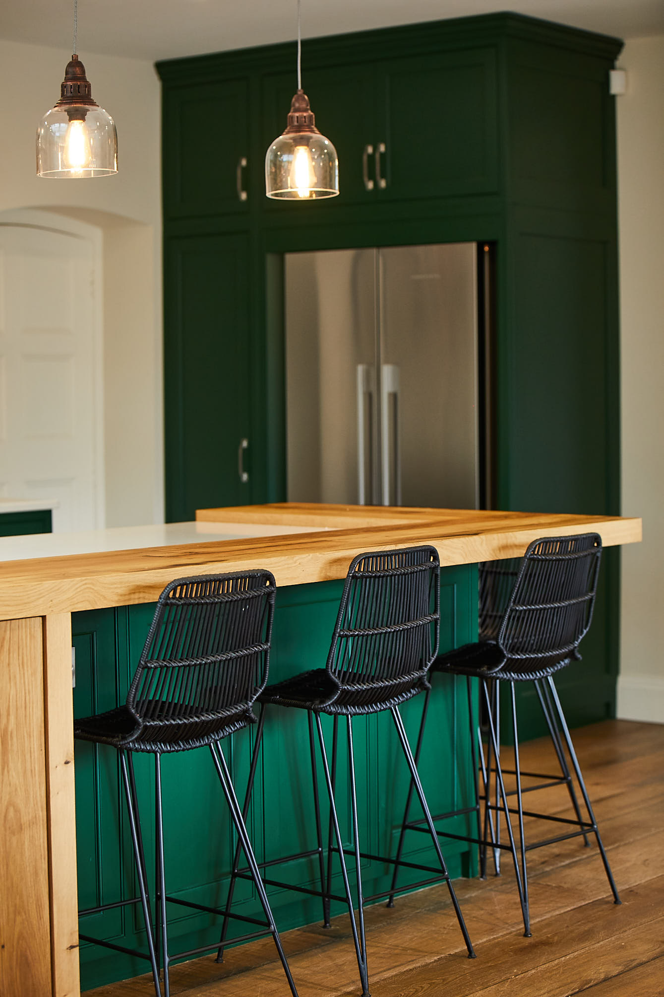 Black bar stools sit under green kitchen island with glass pendant lights above