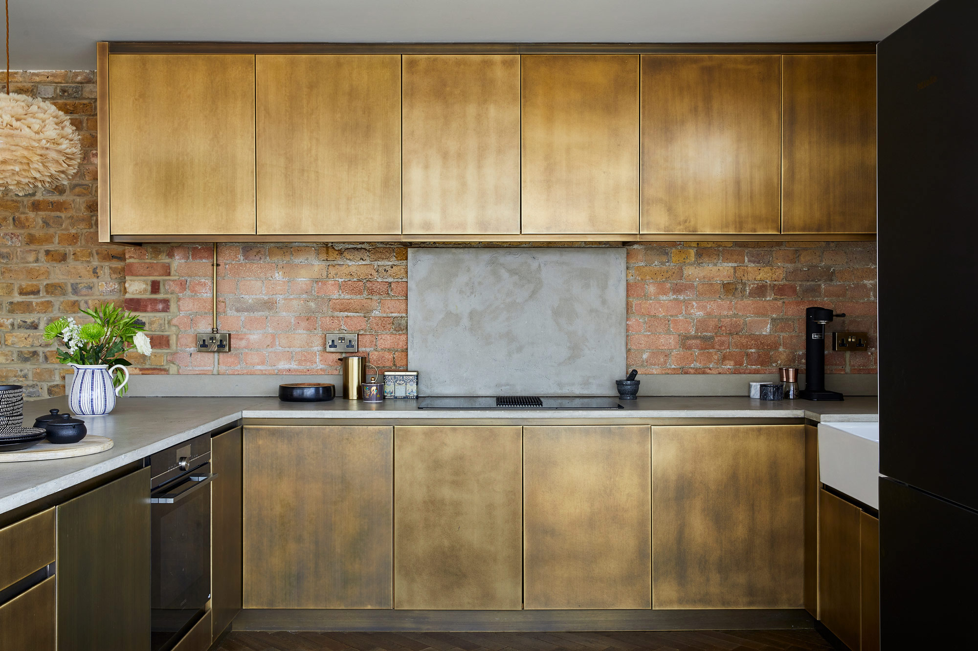 Aged brass kitchen with concrete worktops and exposed brick wall