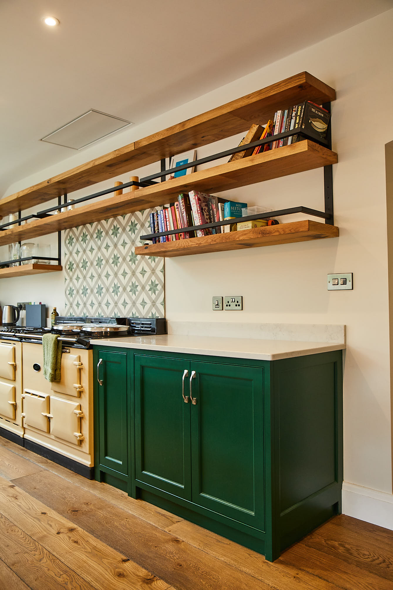 Chunky oak open shelves hang above green kitchen cabinets and cream AGA